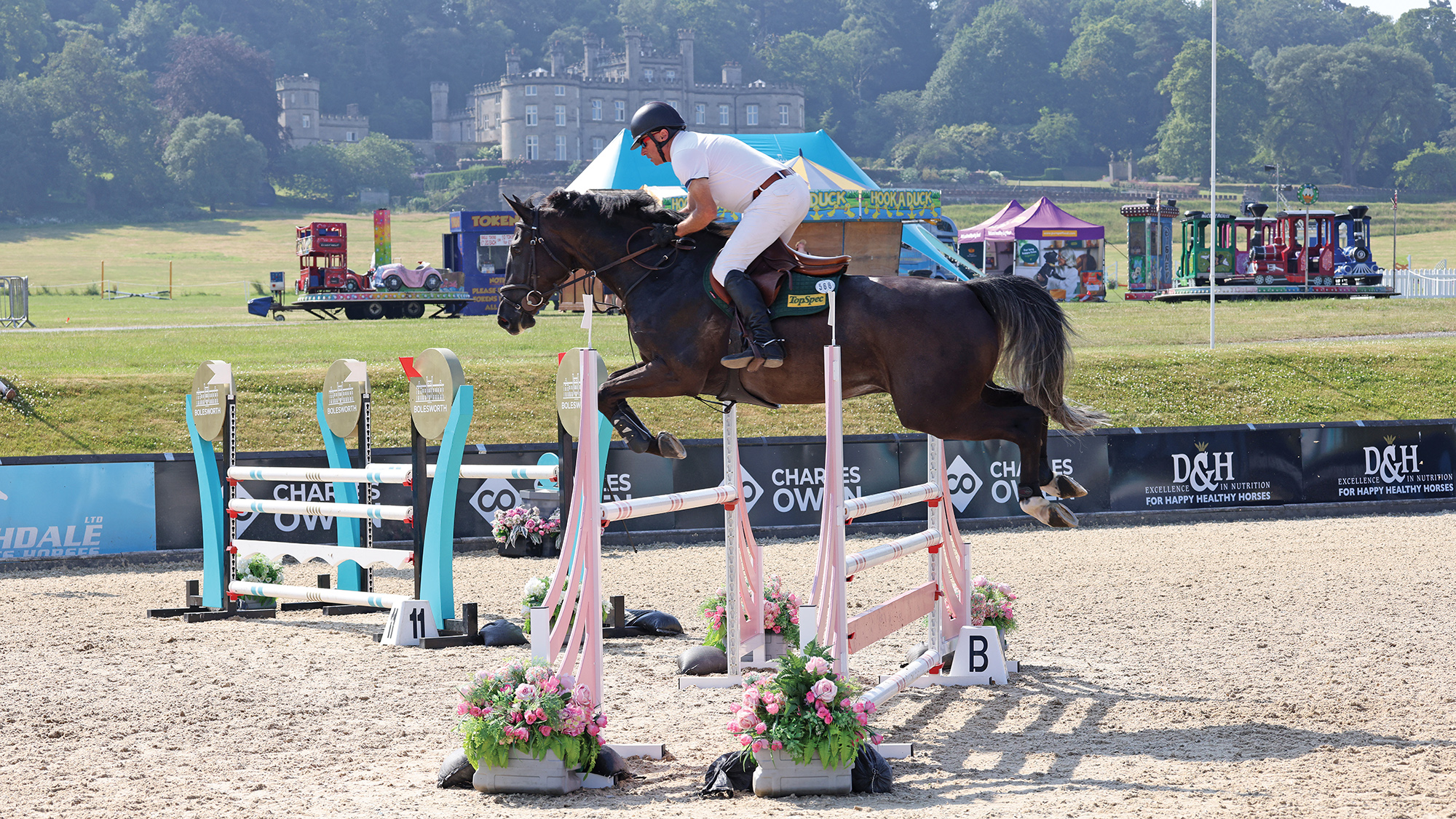 A dark coloured horse jumps a large oxer on a sand arena with Bolesworth Castle in the background.