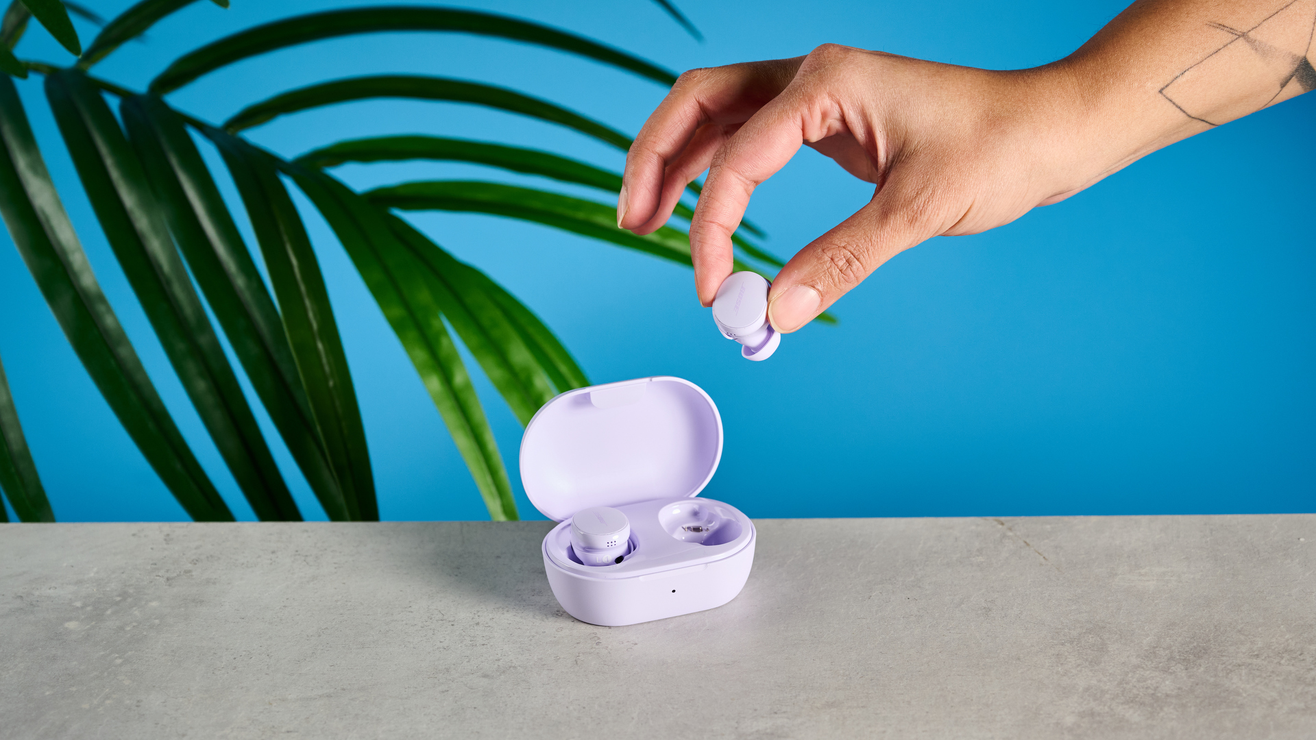 the bose quietcomfort earbuds on a gray table with a purple charging case with a hand taking one of the earbuds out of the case