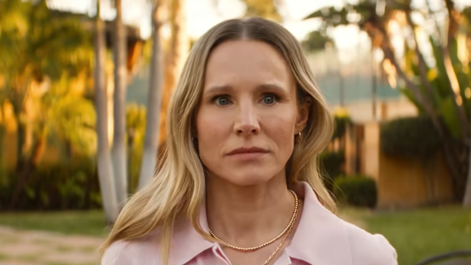 Kristen Bell looking stunned, wearing a pink shirt and standing in front of palm trees in Nobody Wants This Season 3.