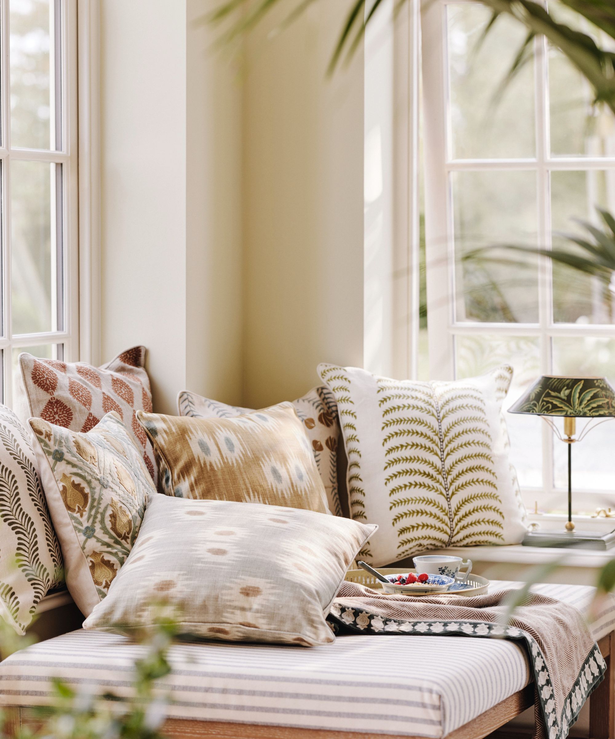 A sunlit daybed piled with layered botanical and ikat print cushions in warm neutrals beside tall white-framed windows with garden views and tropical foliage.
