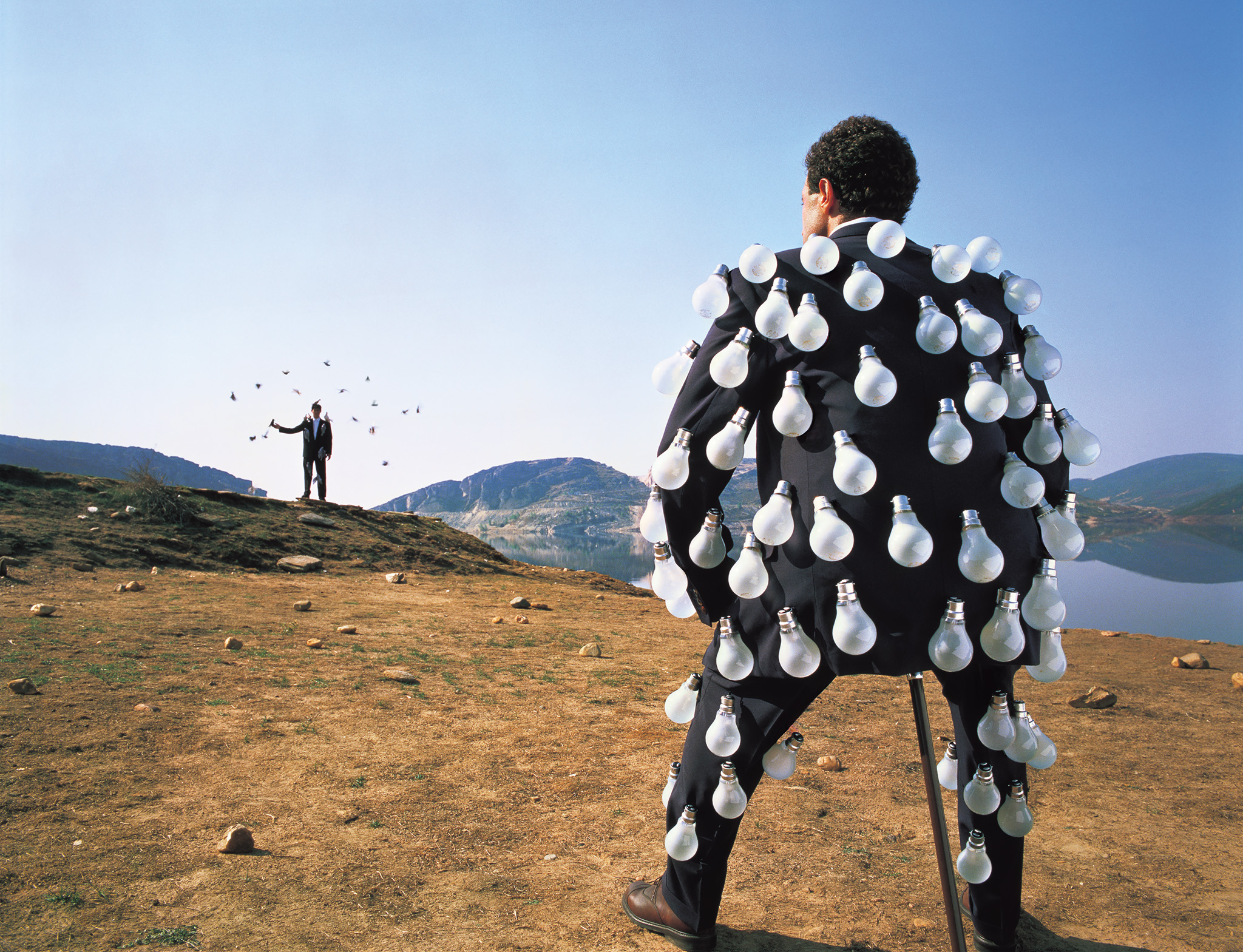 A man wearing a suit covered in light bulbs stands on a barren hillside with his back to the camera, while another man stands further in the distance with birds flying around him.