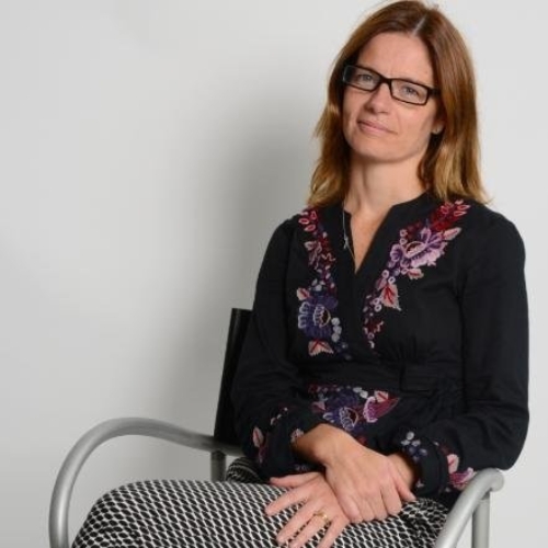 Image of a woman in a black top and black and white pants sitting in a chair in front of a white background.