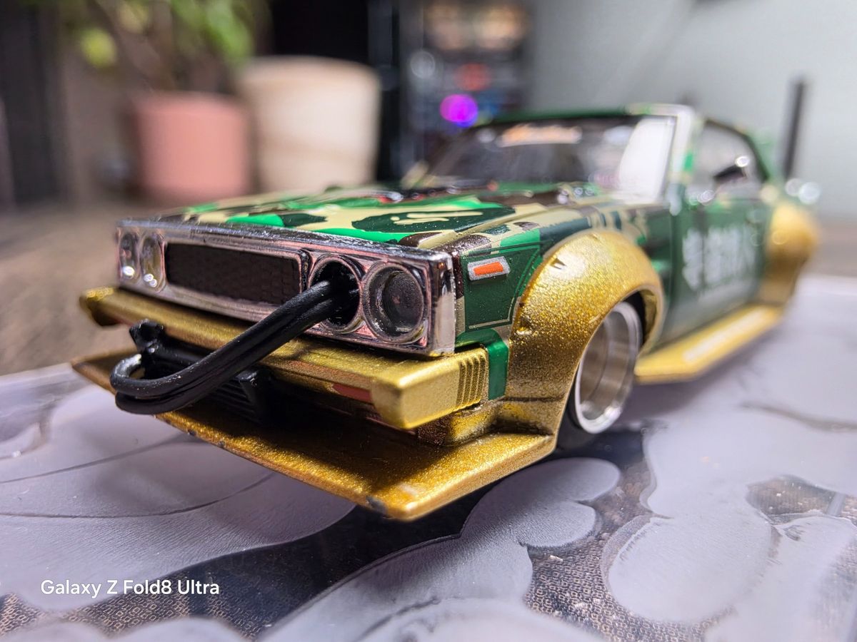 Macro-style close-up of the front of a green and gold model car with camouflage detailing and shallow depth of field.