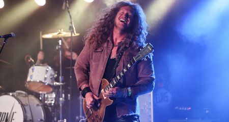 Jared James Nichols smiles as he plays a Les Paul Goldtop live, the stage behind him lit in blue and white.