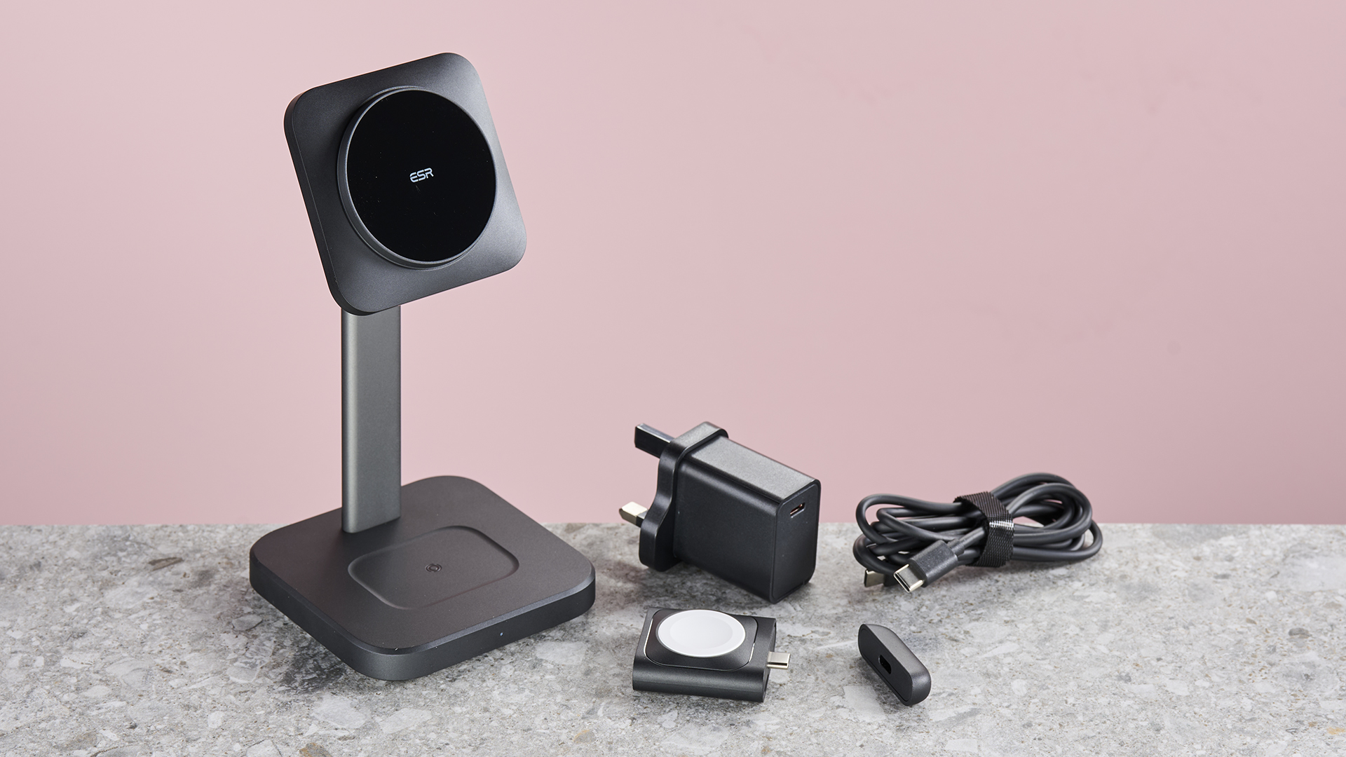 ESR QI2 3 -in -1 Observation Wireless charging kit is sitting on the surface of the light gray stone on the right and leaning on a pink background. On the surface on the right, the plug adapter, the USB-C charging cable and the watch charging component, the cover is unloaded.