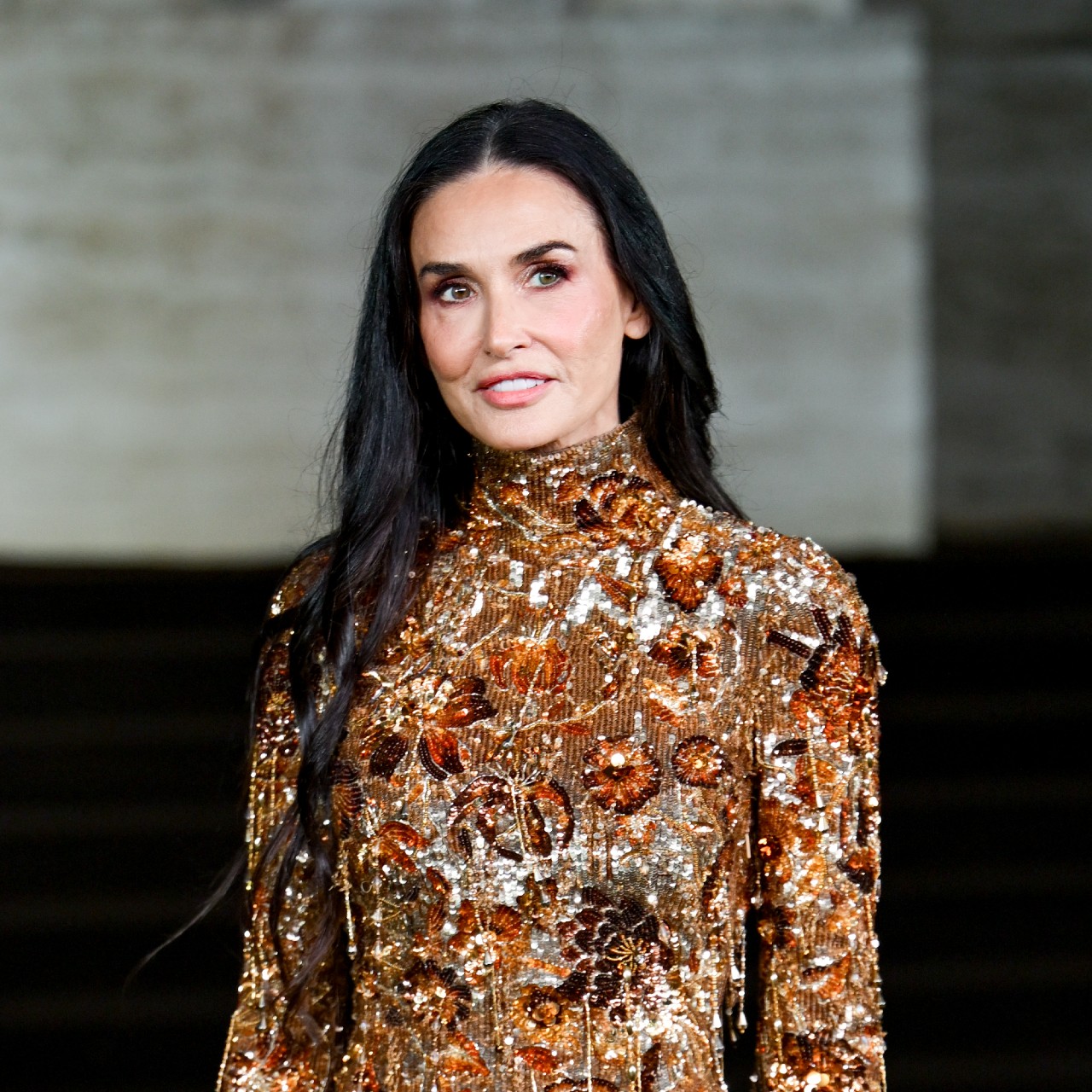a white woman with long dark hair wearing a long-sleeve gold gown with beads and sequins