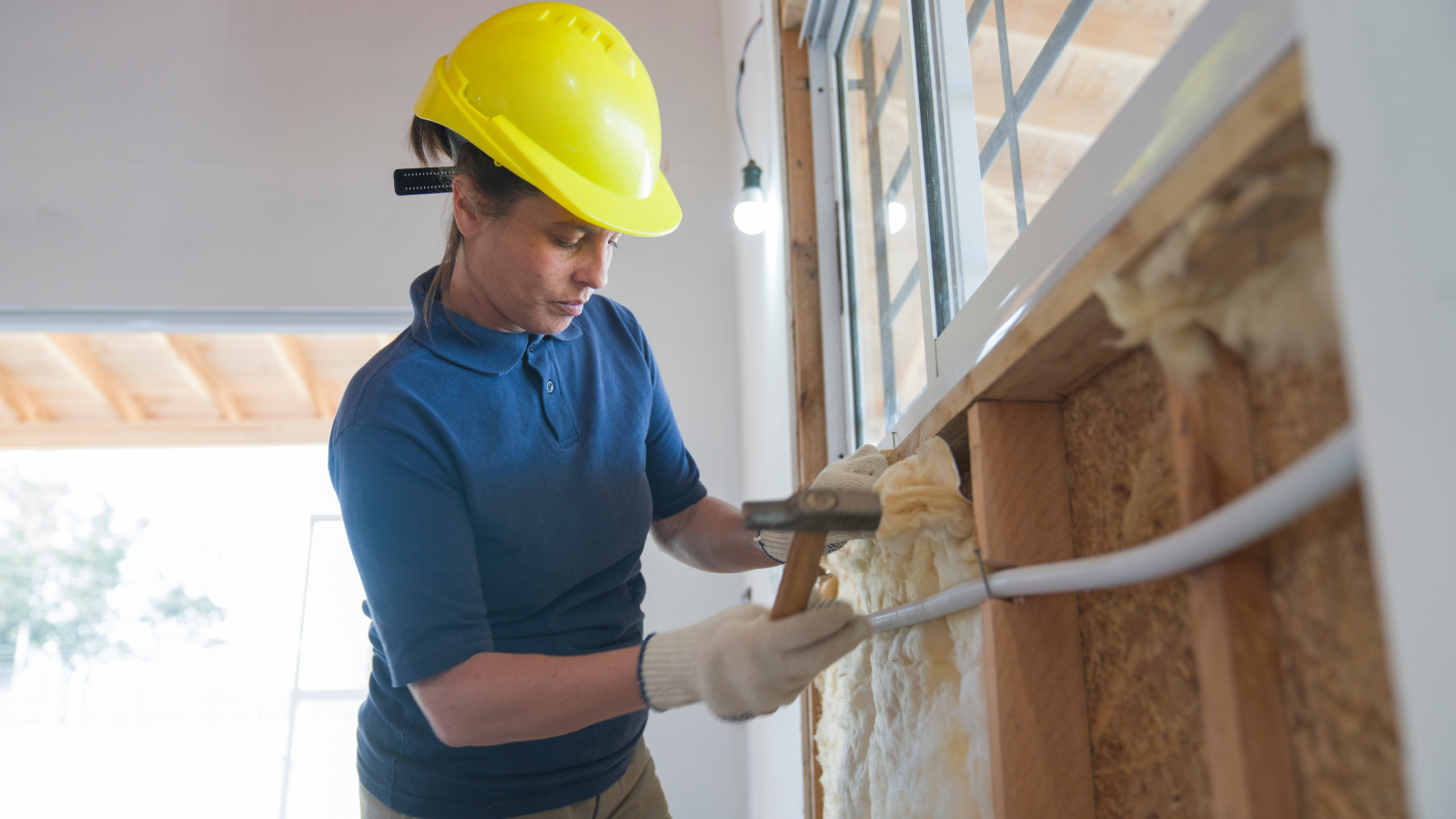 female wearing blue polo shirt, yellow hard hard hat and gloves, inserting insulation into wall