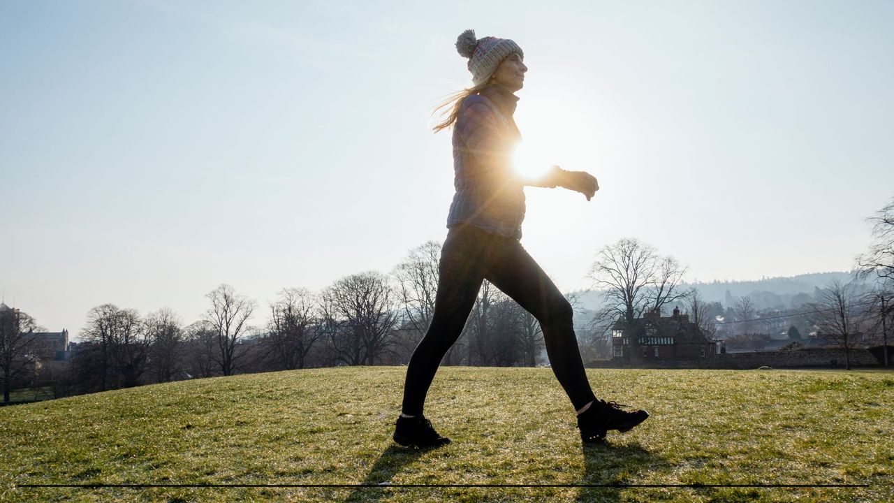 Woman walking briskly across a grass hill, wearing beanie hat and activewear, early morning with sun coming through the clouds