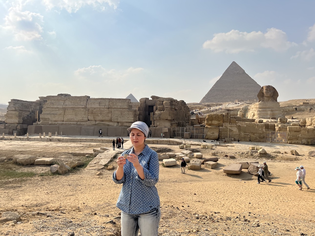 A woman stands in the foreground wearing jeans and a blue shirt with white polka dots. She holds a piece of rock and is looking at it. A pyramid, some stone structures and the Great Sphynx of Giza are all in the background