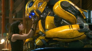 Still from the movie Bumblebee (2018). Here we see the yellow Transformer Bumblebee kneeling in front of a young woman with long dark hair as she takes his face into her hands.