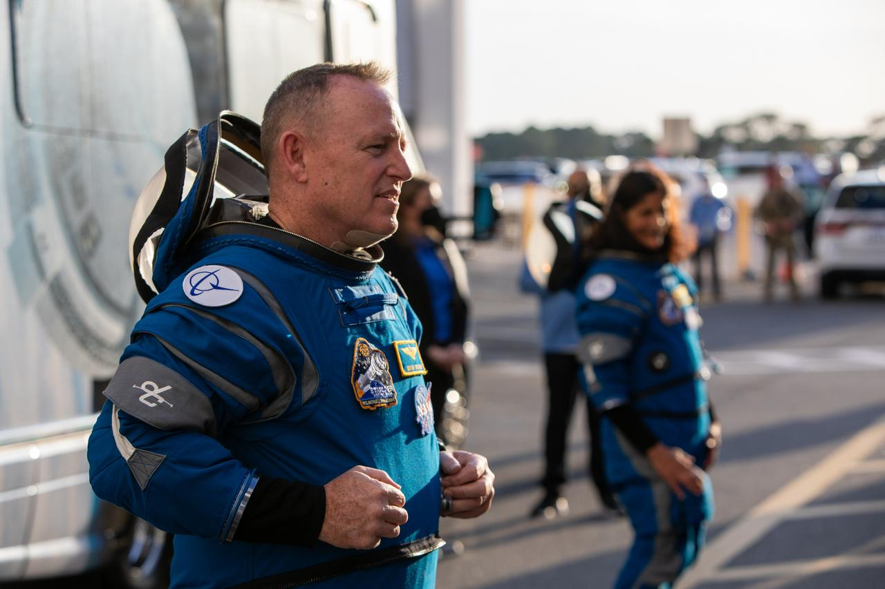 a man with short hair stands on the left, facing right, wearing a blue spacesuit, with the helmet unzipped around the collar and draped back like a hoodie. Another astronaut, out of focus, stands to his side, upward and to the right in the image. Par of a bus can be seen behind them, and crowds standing at the edge of a parking lot in the background.