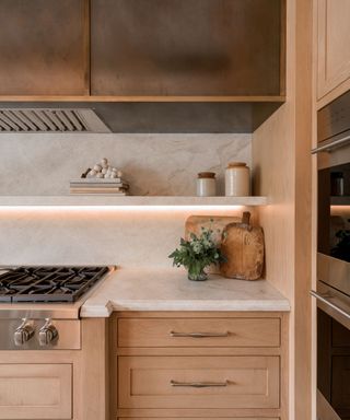 A wooden kitchen with natural warm white stone countertops and backsplash. A stone floating shelf blends into the backsplash and topped with decor, with under-shelf lighting to add extra illumination