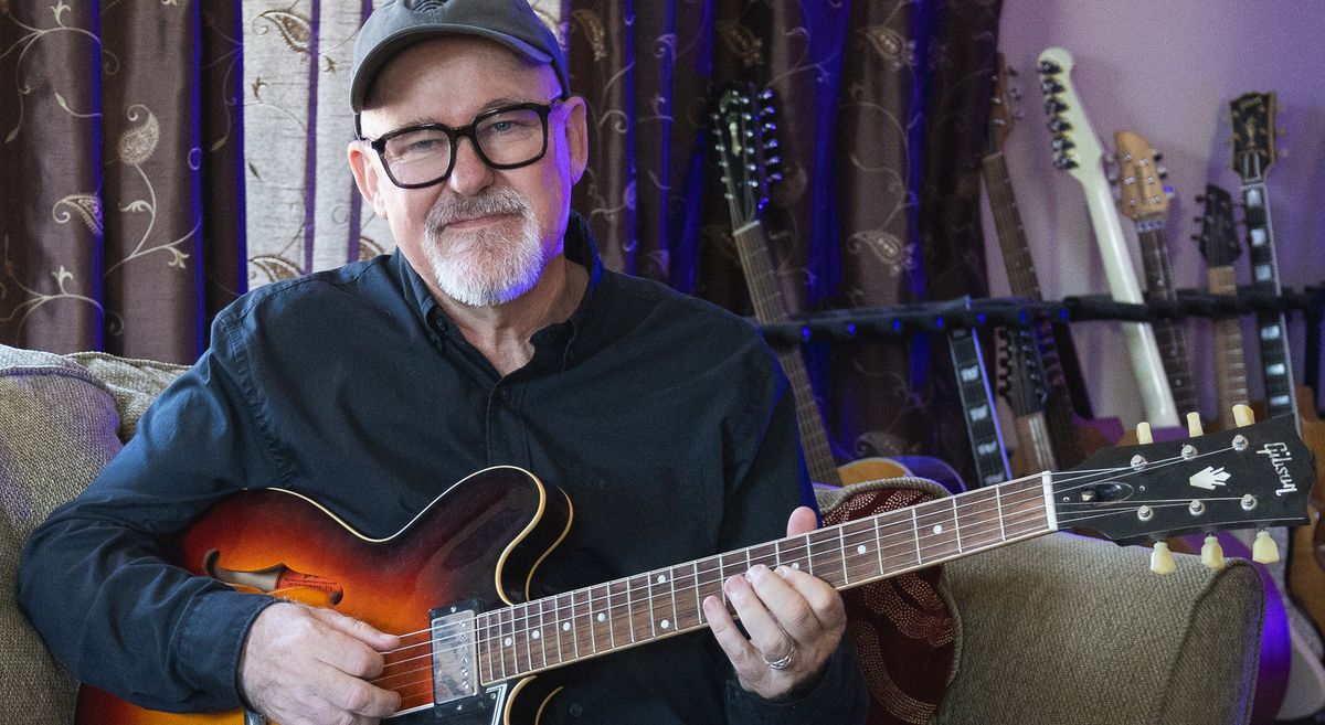 Session pro Tim Pierce is photographed at home, wearing a ballcap and fretting a chord on his guitar.