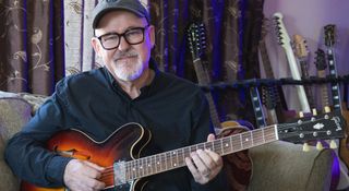 Session pro Tim Pierce is photographed at home, wearing a ballcap and fretting a chord on his guitar.