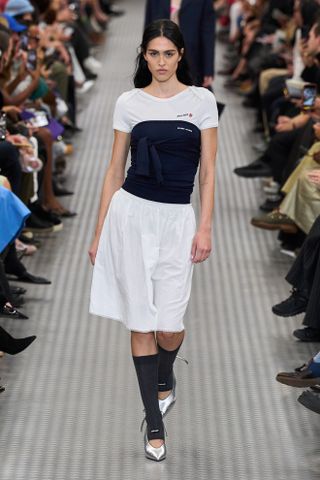 Amelia Hamlin wearing a white Miu Miu T-shirt with a strapless navy blue top, white midi skirt, socks, and silver heels at the Miu Miu spring summer 2025 runway show in Paris.