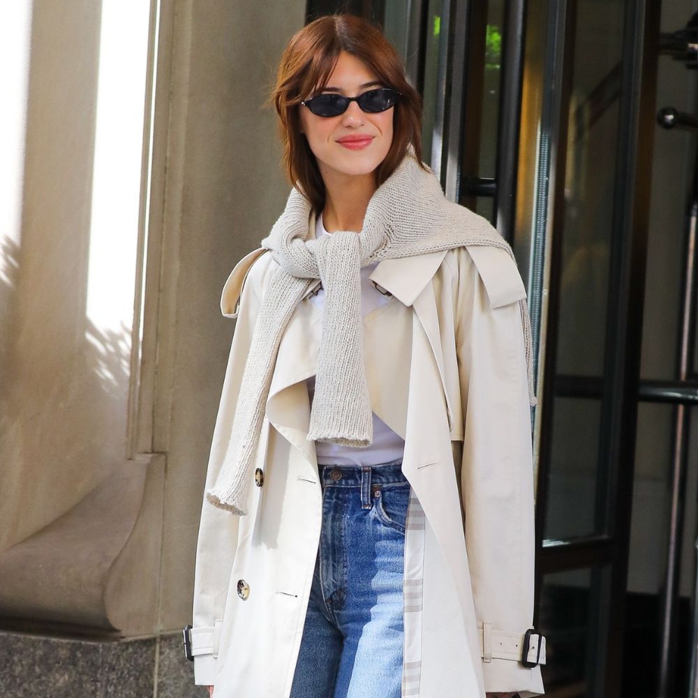 Daisy Edgar Jones steps outside of a hotel wearing a beige trench coat with high-waisted stovepipe jeans, black kitten heel boots and a beige knit over her shoulders.