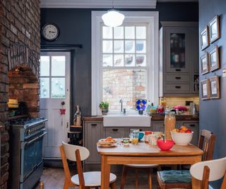 kitchen painted in blue with exposed brick wall housing range cooker, wooden table and chairs and butler sink in front of white sash window