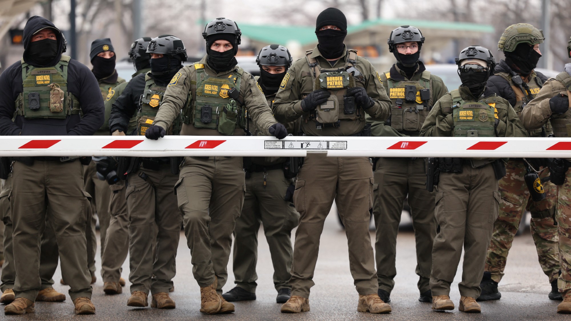 a row of uniformed ICE officers standing in front of a barricade. they are all masked. 