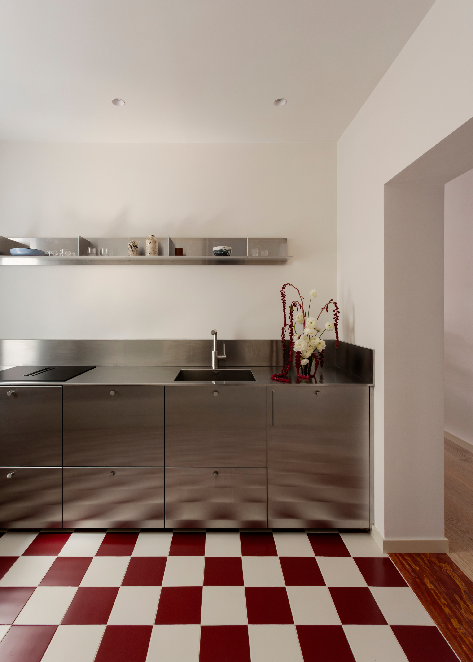 A white kitchen with cool chrom floating shelves, and a chrome workspace with checkerboard tiled flooring, and a vase of roses and red stems