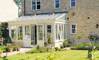 a traditional conservatory/orangery with white painted windows and door from David Salisbury added onto a light brick house, with a pretty garden full of plants and flowers outside