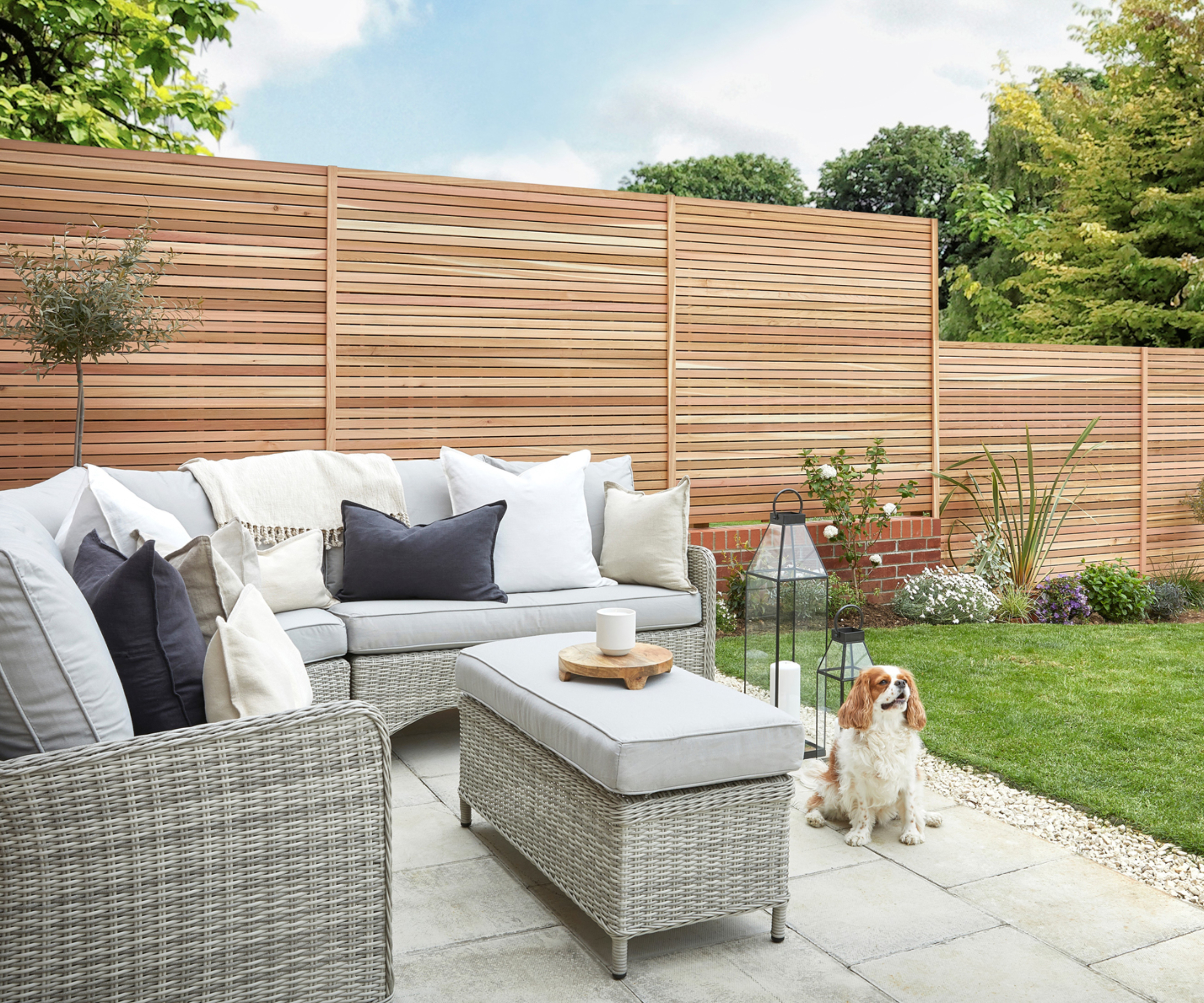 modern slatted fence in garden with paved patio, rattan furniture, lawn and small dog sat on patio