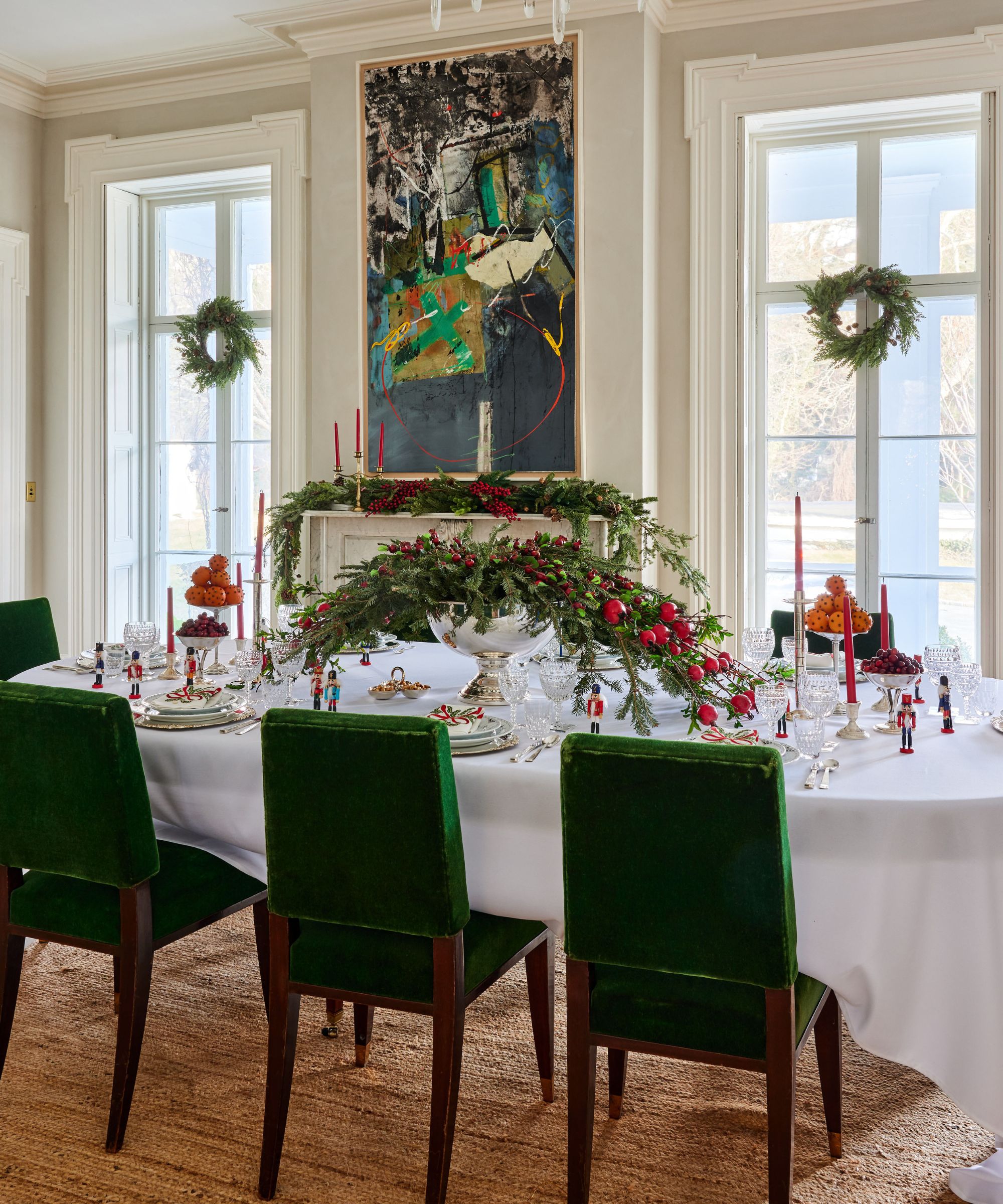 A traditional dining room with large windows and period features. A large dining table with a white tablecloth and green velvet dining chairs, with festive foliage decor.