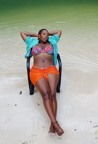 A woman sitting in a chair by the beach wearing a floral bikini with an orange sarong and floral print button-down shirt.