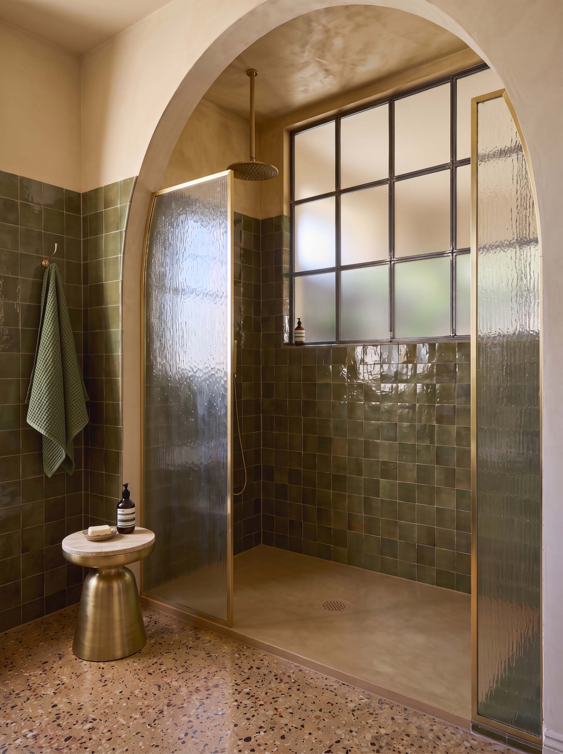 Warm beige shower tray with olive-green wall tiles, brass-framed glass screens, and a pink terrazzo floor beneath a plaster arch.