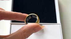 Thumb and index finger hold a gold Oura ring. Smal sensors can just be seen inside the band