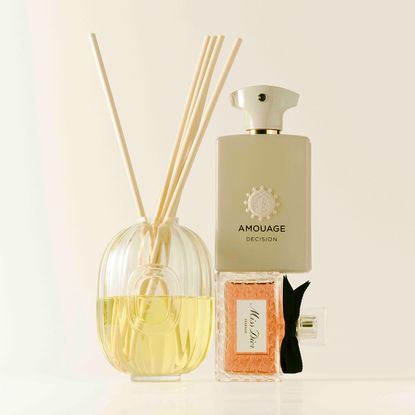 diptyque diffuser next to a miss dior fragrance bottle with an amouage decision fragrance bottle on top, on a cream colored background