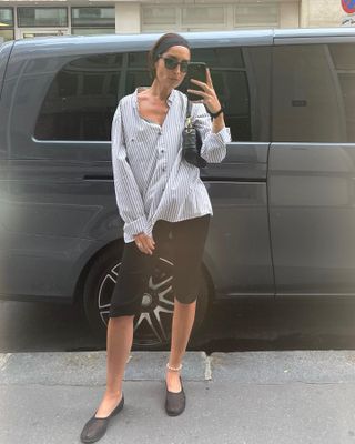 influencer Géraldine Boublil poses for a window-reflection selfie wearing a black headband, striped button-down shirt, designer shoulder bag, black capri pants, a pearl anklet, and black mesh flats