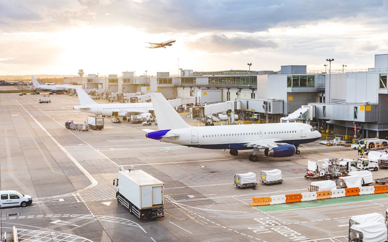 Busy airport view with airplanes and service vehicles at sunset. London airport with aircrafts at gates and taking off, trucks all around and sun setting on background. Travel and industry co