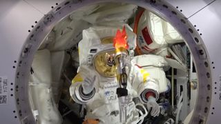 an astronaut in a white spacesuit prepares to catch a simulated olympic torch inside a white-walled space station module