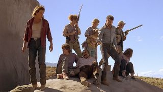 A scene from the movie Tremors (1999). Here we see a group of 9 people standing on a large boulder. They are looking wearily round them, two of them holding rifles at the ready.