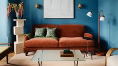 A terracotta sofa in a blue living room. There is a glass coffee table in front of the sofa and a terracotta floor lamp on the right side of the sofa. On the left, is a cream, structure that has a flower vase on top of it.