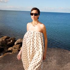 A female model wearing a black and white polka dot dress and black sunglasses standing on a rock with the sea behind her