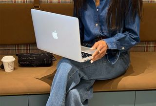 Woman with a laptop on her lap, wearing a button down denim shirt and light washed jeans.