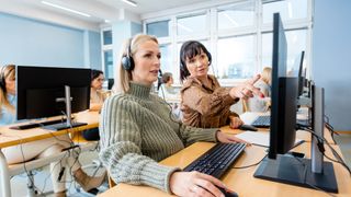 Two female IT helpdesk staff collaborating on desktop computers while working in an open plan office space.