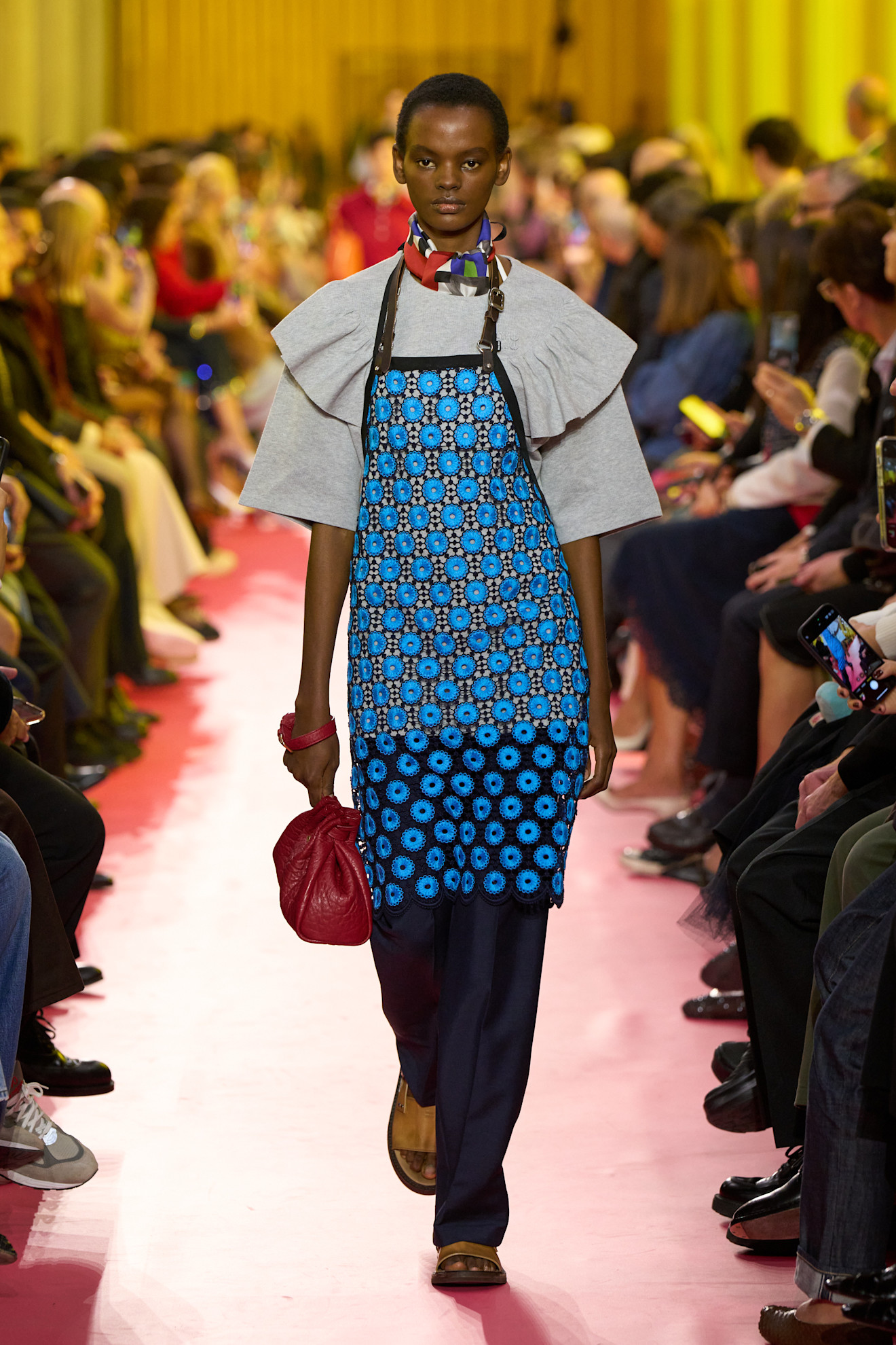 a model walking in Miu Miu Sprin 2026 wearing a blue apron, grey top, multicolored scarf, blue pants, tan slides, and a red bag