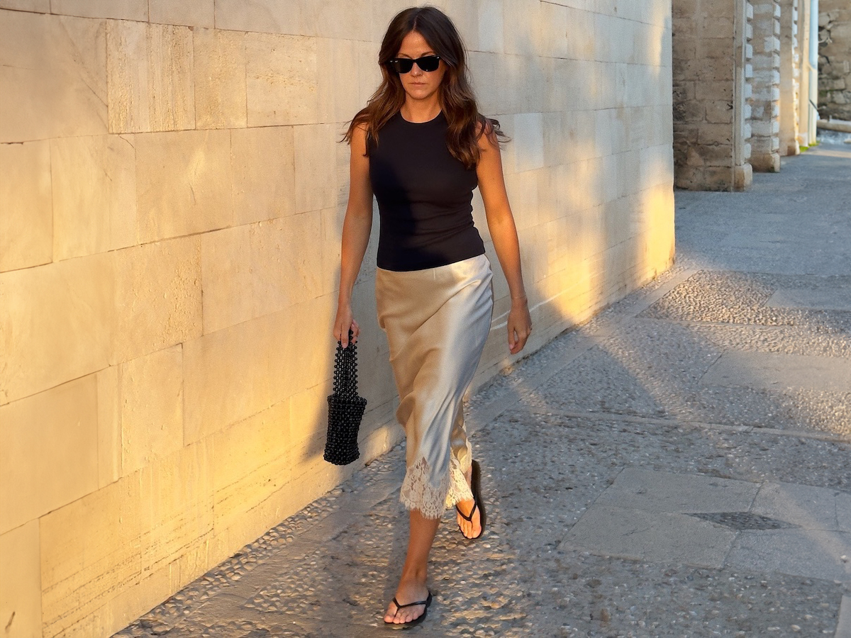 British influencer Kim Turkington walks in the golden hour of Palma de Mallorca, Spain wears chic summer outfit ideas, black sunglasses, black vests, silk ivory skirt with lace hem, black beaded handbag and black flat-plane trailer sandals.