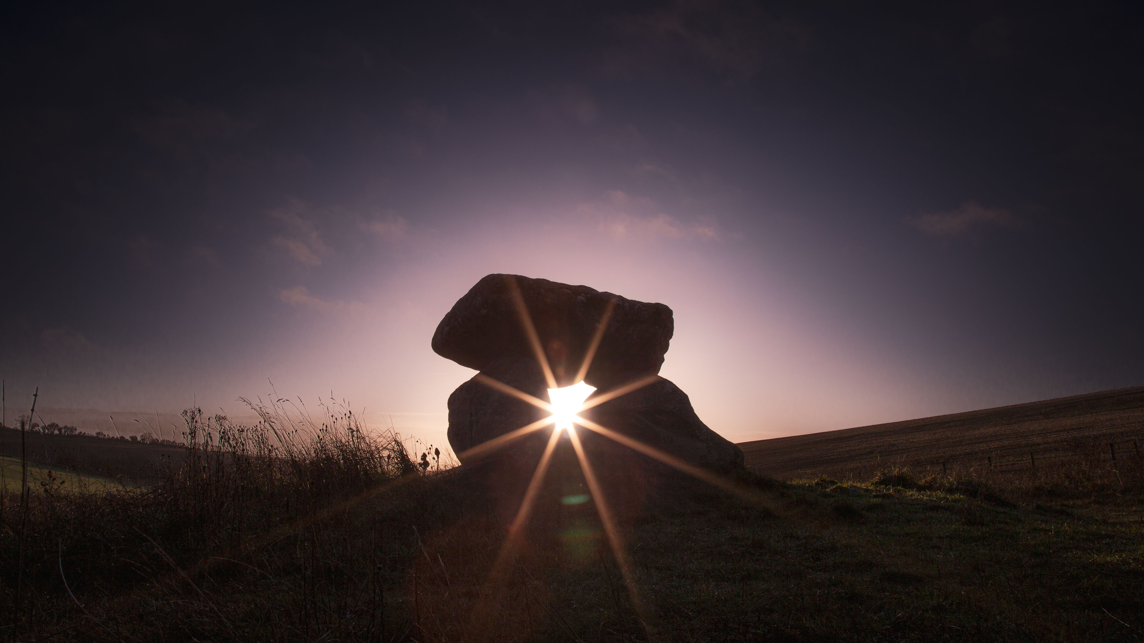 the sun rises in between a pillar of stones grass is visible on the left in shadow, backdropped against the sky, and a hill rises to the right