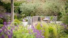 A garden with a flower bed in the foreground. The camera is focussed on an outdoor table and chairs on a patio area