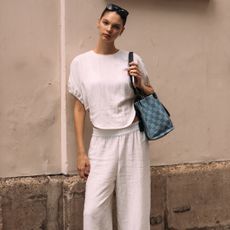 woman wearing white linen set, checkered bag, and sunglasses standing in front of concrete wall
