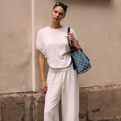 woman wearing white linen set, checkered bag, and sunglasses standing in front of concrete wall
