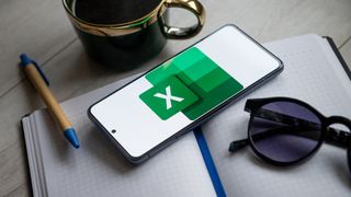 Microsoft Excel logo and branding pictured on a smartphone with a notepad and coffee cup.