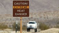 A shimmering desert road is hot with a brown sign in the front reading "Caution, Extreme heat Danger." with a blurred car in the background