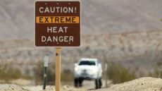A shimmering desert road is hot with a brown sign in the front reading "Caution, Extreme heat Danger." with a blurred car in the background