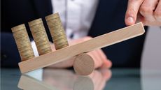 Businessman balances stacked coins with finger on wooden seesaw over reflective desk.