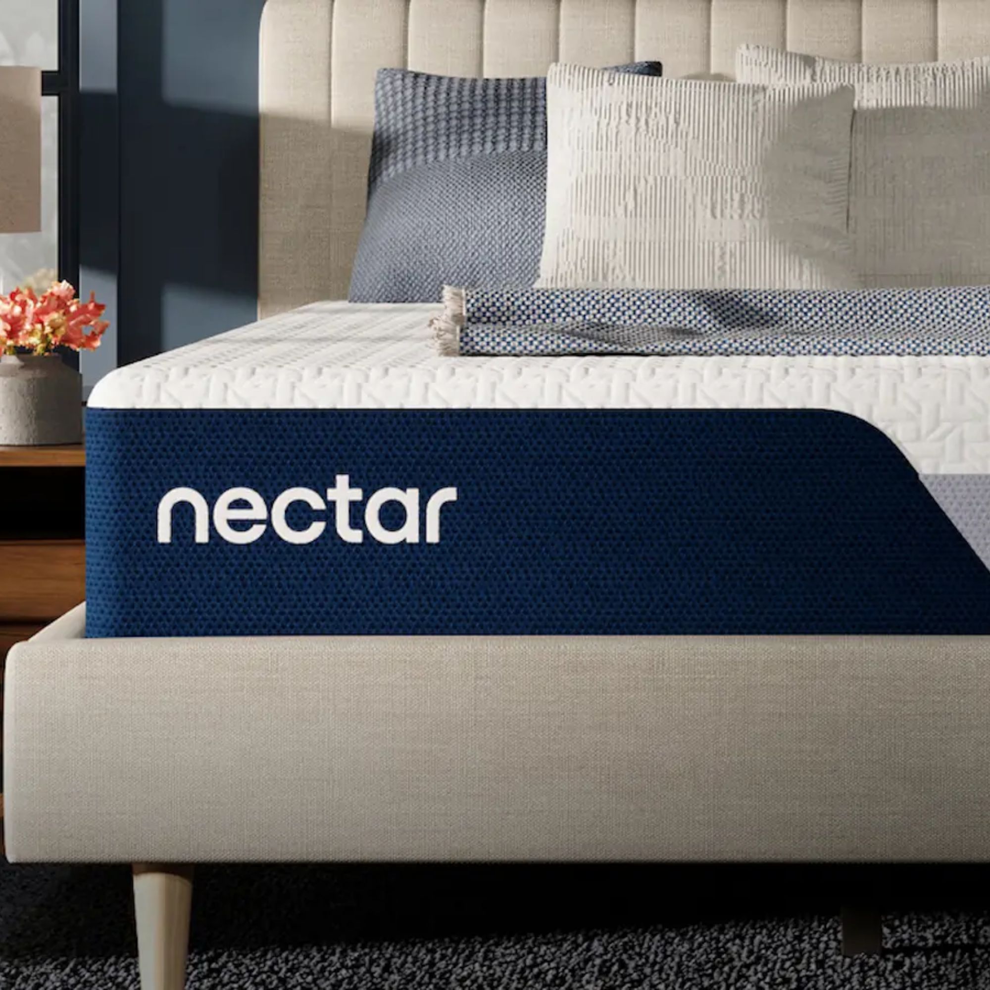 A white Nectar Classic Memory Foam Mattress with a blue edge on a neutral upholstered bed frame with a scalloped headboard. On top of the bed are blue and white cushions and a blue and white folded blanket. To the left of the bed is a dark blue wall and a wooden side table with a grey vase with orange flowers in it. The carpet beneath the bed is dark grey.