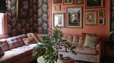 A living room with floral wallpaper, a gallery wall, a dusky pink L-shaped couch, and a marble table with a plant and books on top of it